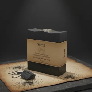 A dark grey to black bar of Kemit natural Activated Charcoal Soap, inspired by ancient Egyptians, is displayed on a piece of aged brown paper with an Eye of Horus illustration. The paper and soap rest on a dark grey stone slab, with loose charcoal dust and a small chunk of charcoal scattered around.