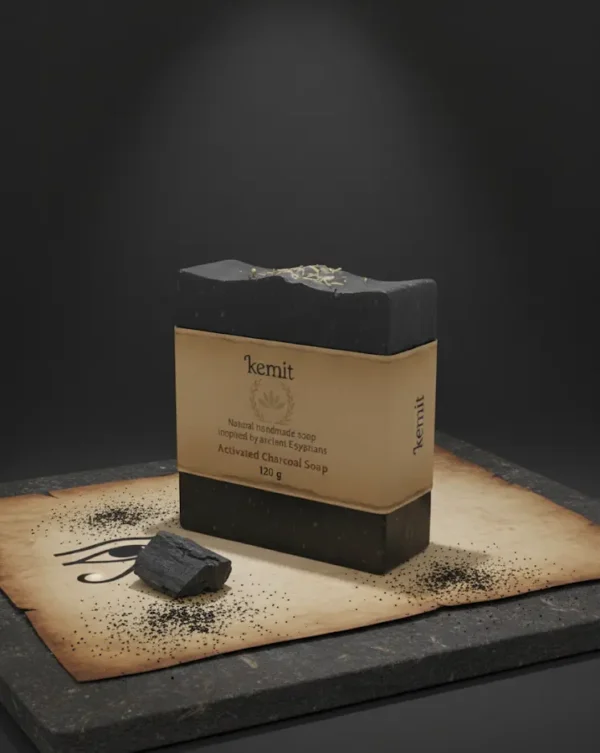 A dark grey to black bar of Kemit natural Activated Charcoal Soap, inspired by ancient Egyptians, is displayed on a piece of aged brown paper with an Eye of Horus illustration. The paper and soap rest on a dark grey stone slab, with loose charcoal dust and a small chunk of charcoal scattered around.