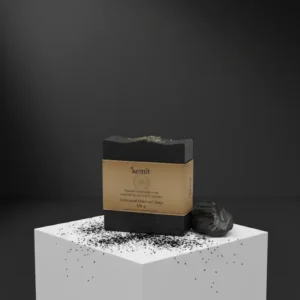 A dark grey to black bar of Kemit natural Activated Charcoal Soap, inspired by ancient Egyptians, sits on a white cube pedestal. A chunk of charcoal and scattered charcoal dust are visible on the cube's surface. The background is a dark grey, with a spotlight shining from above.