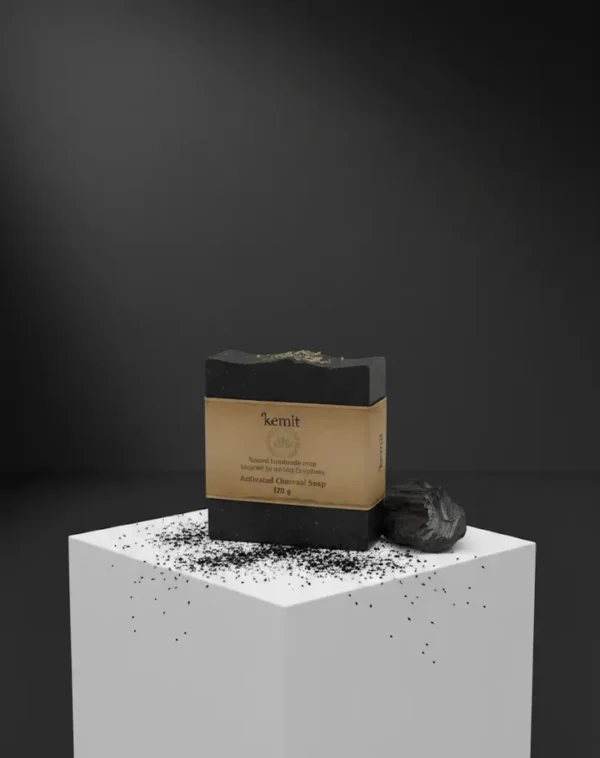 A dark grey to black bar of Kemit natural Activated Charcoal Soap, inspired by ancient Egyptians, sits on a white cube pedestal. A chunk of charcoal and scattered charcoal dust are visible on the cube's surface. The background is a dark grey, with a spotlight shining from above.