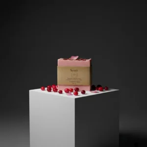 A reddish-pink bar of Kemit natural Cranberry Soap rests on a white cube pedestal, surrounded by scattered fresh cranberries and a few rose petals. A small amount of dried petals and fruit bits are visible on top of the soap bar. The background is a dark grey.