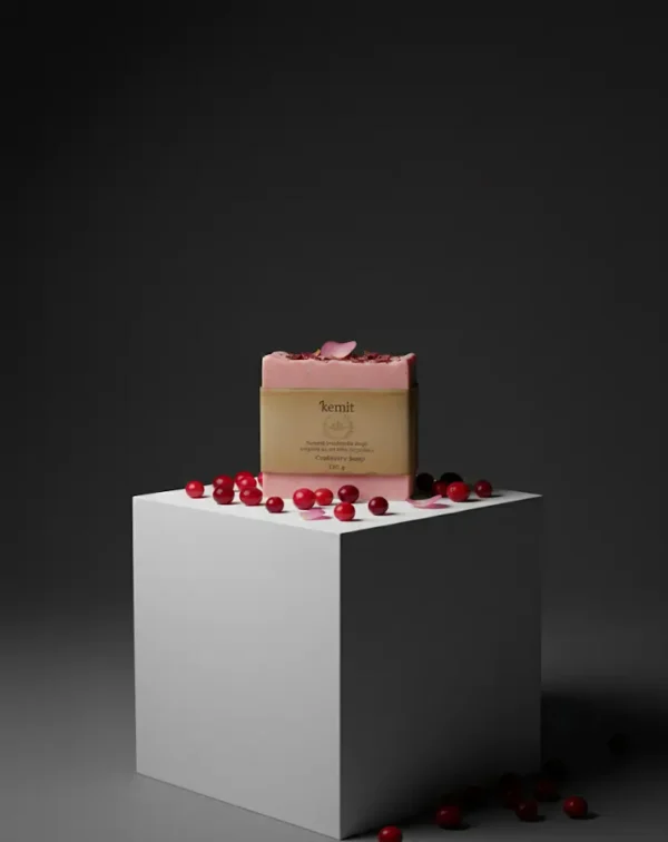 A reddish-pink bar of Kemit natural Cranberry Soap rests on a white cube pedestal, surrounded by scattered fresh cranberries and a few rose petals. A small amount of dried petals and fruit bits are visible on top of the soap bar. The background is a dark grey.