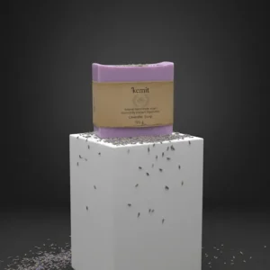 A purple bar of Kemit natural handmade Lavender Soap sits on a white cube. Dried lavender buds are scattered across the top of the cube and on the dark surface below.