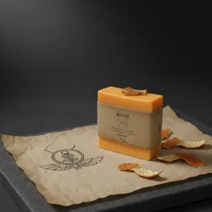A vibrant orange bar of Kemit natural Orange Soap, inspired by ancient Egyptians, is displayed on a piece of crumpled brown paper with the Egyptian sun illustration. The paper and soap rest on a dark grey stone slab, with several dried orange peels scattered around. A single dried orange peel sits on top of the soap bar.