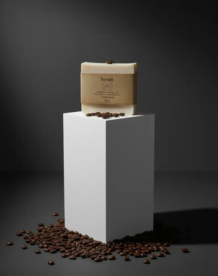 A creamy white bar of Kemit natural Coffee Soap sits on a white cube pedestal. A pile of roasted coffee beans is scattered around the cube's base. A single coffee bean rests on top of the soap bar.
