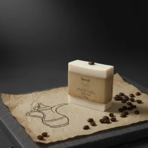 A creamy white bar of Kemit natural Coffee Soap is displayed on a crumpled brown paper with a stylized Arabic coffee pot illustration. The soap is on a grey stone slab, with several coffee beans scattered on the paper and the surface below. A single coffee bean rests on top of the soap.