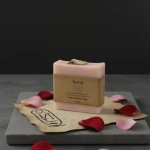 A light pink bar of Kemit natural Rose Balsam Soap, inspired by ancient Egyptians, is displayed on a piece of crumpled brown paper with an Egyptian hieroglyphic-style illustration. The paper and soap rest on a dark grey stone slab, surrounded by scattered pink and red rose petals. A single dried rose petal adorns the top of the soap.