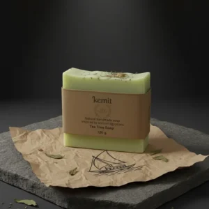 A light green bar of Kemit natural Tea Tree Soap, inspired by ancient Egyptians, is displayed on a piece of crumpled brown paper with a stylized boat illustration. The paper and soap rest on a rough grey stone slab. Dried leaves and small pieces of wood are scattered around the soap.