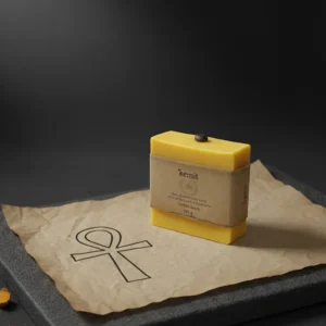 A vibrant yellow bar of Kemit natural, handmade soap is presented on a piece of rustic brown paper with an Ankh symbol drawn on it. A single coffee bean rests on top of the bar, which is wrapped with a simple brown band. The background is a stark black. In the foreground, there are scattered grains of coffee and pieces of turmeric root.
